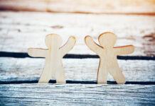 Reimaginar el Apoyo en Crisis, conversación con Tina Minkowitz. Silueta de dos personas, hechas con madera, hacen el ademán de tomarse de las manos en gesto de apoyo y acompañamiento.